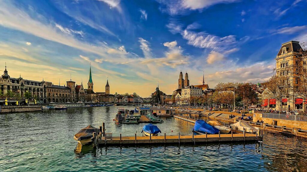 A scenic view of a European city with historic buildings lining a river, a small dock with boats, and a vibrant sunset sky while travelling to Switzerland in September.