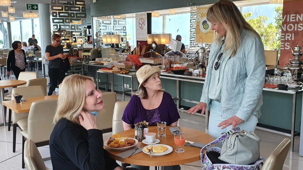 Three women conversing in a busy cafeteria with people in the background and a food counter laden with various dishes, guided by their Trafalgar travel director.