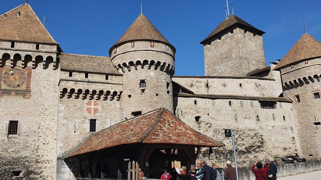 Medieval castle with fortified walls and round towers, featuring a large clock, under a clear blue sky. Trafalgar travel director and tourists are visible near the entrance.