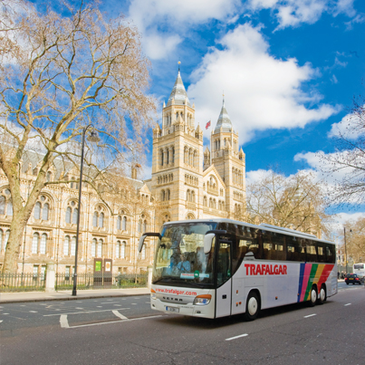A coach travels on a sunny street with the natural history museum in the background, under a clear blue sky.
