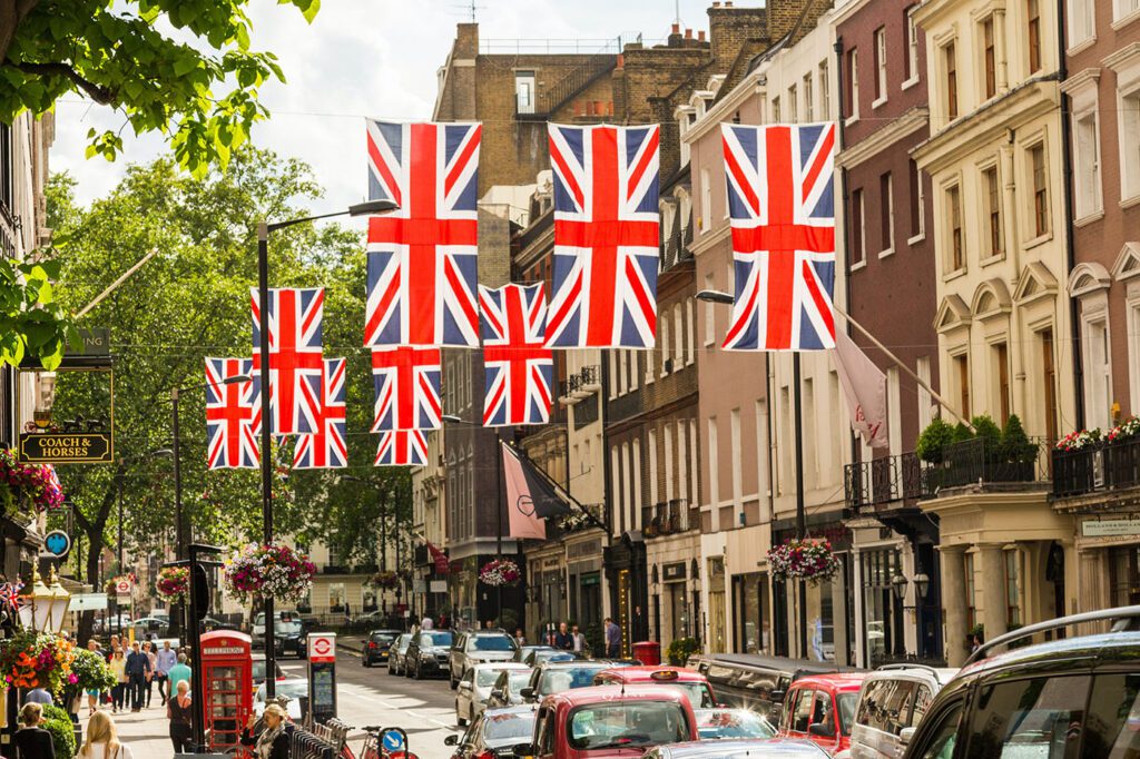 Street decorated with quintessentially British union jack flags in a city, with cars and pedestrians visible, suggesting a festive or national celebration.