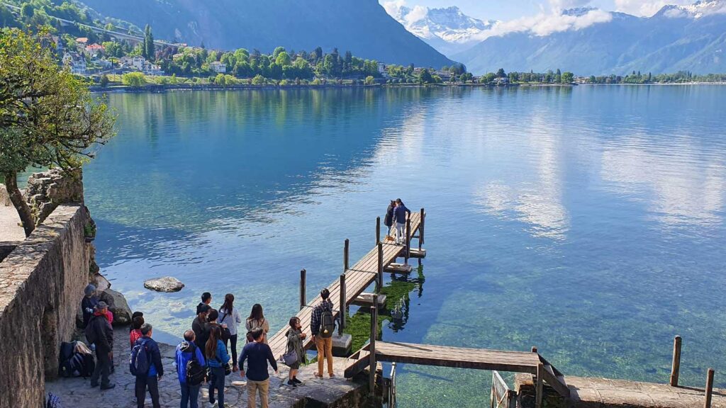 People on a wooden pier at a lake with mountains in the background, under a clear sky. Some individuals, guided by their Trafalgar travel director, are preparing to dive into the water.