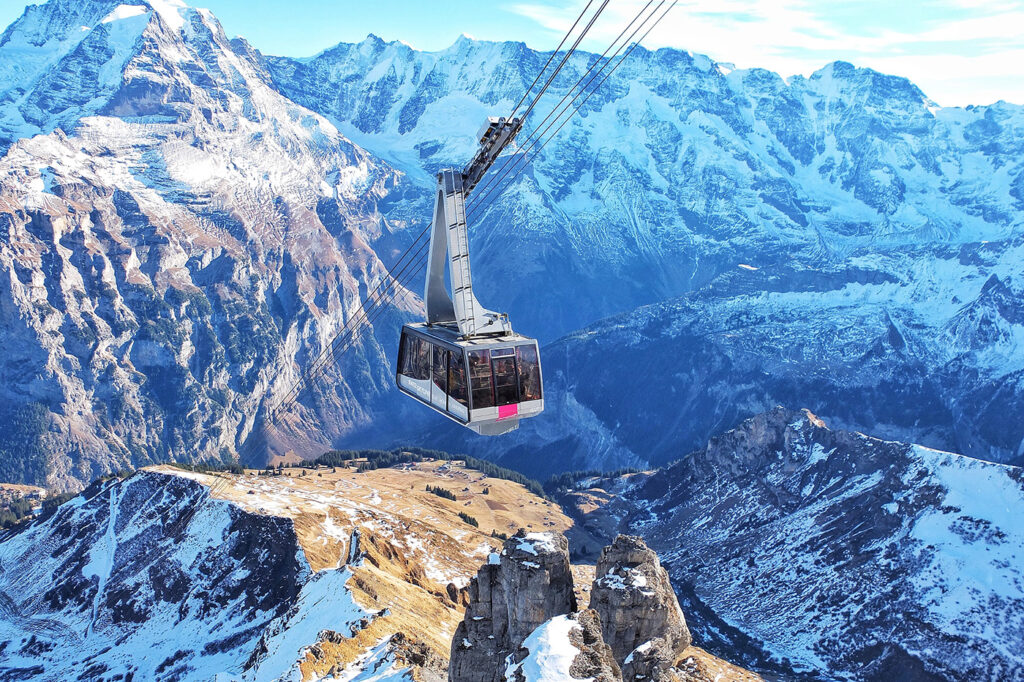 Cable car over mountainous terrain with snow-covered peaks in the background and rocky foreground under a clear blue sky, ideal for sports tours.