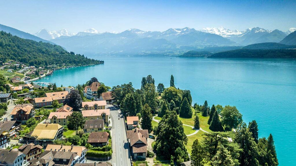 Aerial view of a lakeside village in Switzerland with turquoise water, surrounded by a lush green landscape and distant snow-capped mountains.