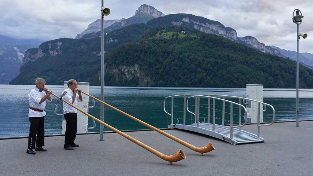 Two elderly men play long alphorns on a lakeside dock with mountains in the background under an overcast sky, while travelling to Switzerland in September.