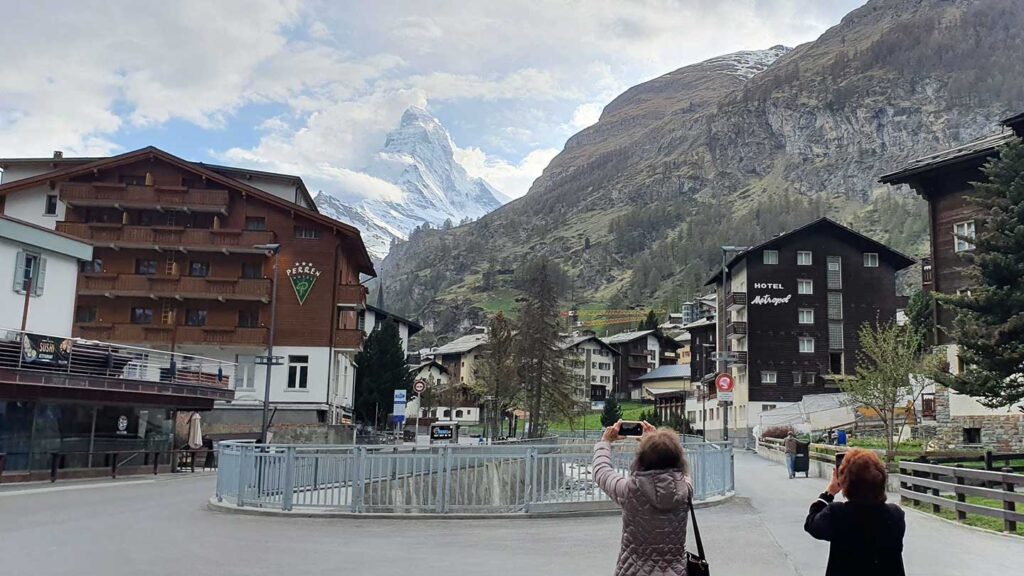 Two people taking photos of the Matterhorn mountain visible behind a quaint village with buildings and a clear sky, accompanied by their Trafalgar travel director.