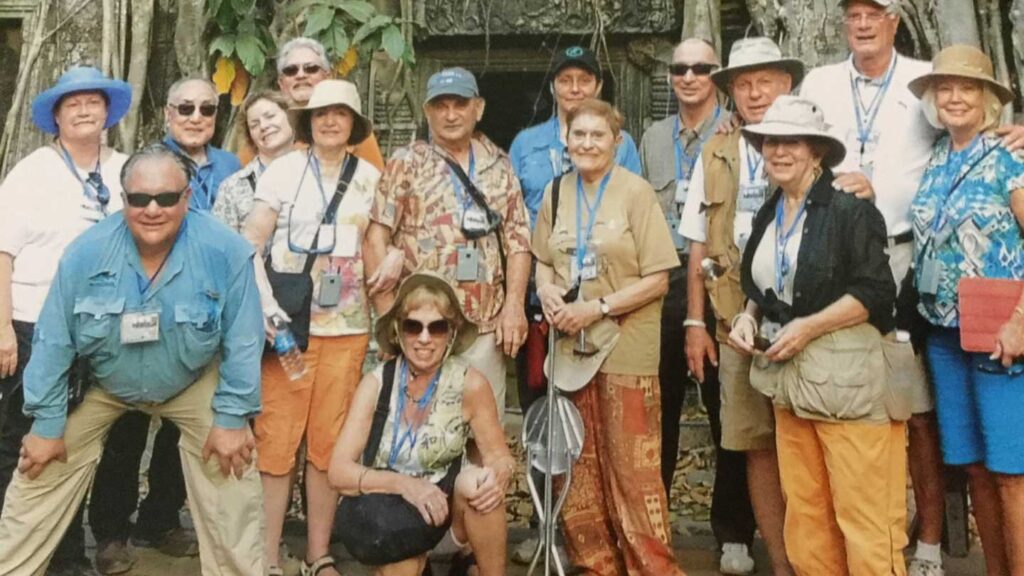 Group of elderly Trafalgar guests posing for a photo in a tropical forest location, wearing casual clothing and hats, some holding cameras.