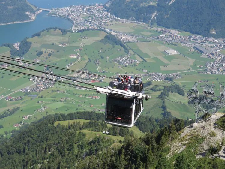A cable car filled with passengers ascends over a scenic landscape featuring a valley with green fields and a small town near a lake, highlighting reasons to travel to Switzerland.
