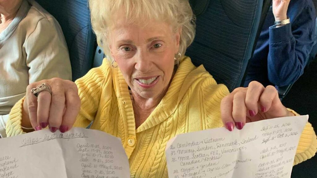 Elderly Trafalgar guest in a yellow sweater happily displaying two handwritten pages on an airplane.