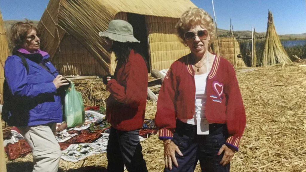 Three women, one in a red jacket, the other two conversing, standing on a reed island with thatched structures and a Trafalgar guest in the background.