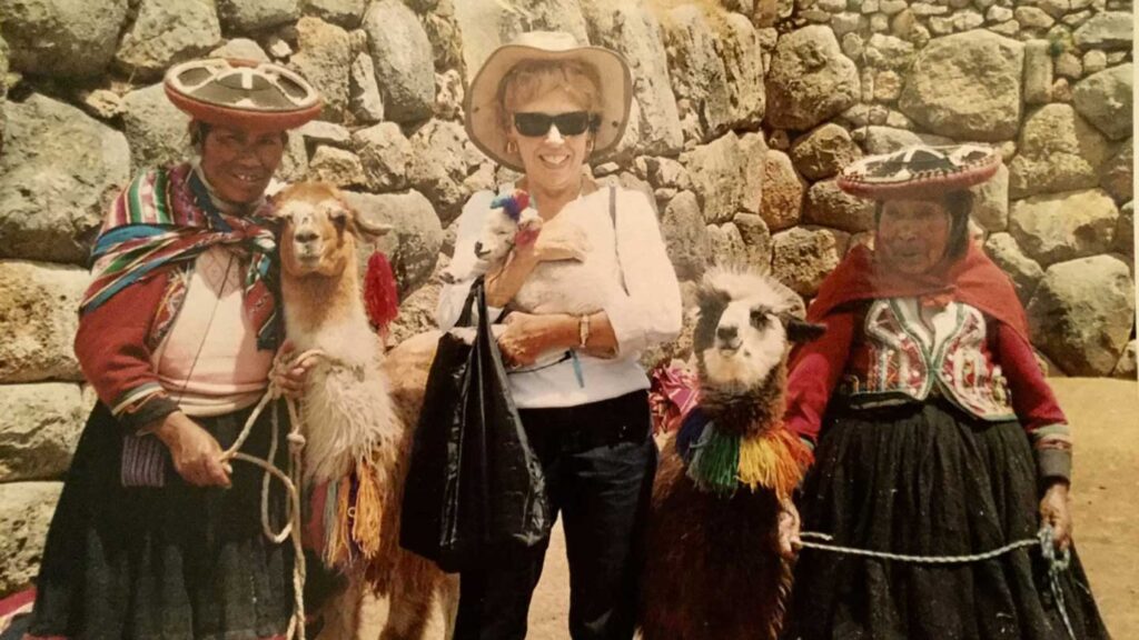 A Trafalgar guest in a sunhat poses with two local women in traditional Peruvian attire and adorned llamas in front of ancient stone walls.