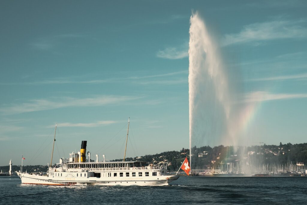 Vintage paddle steamer cruising on a lake with a large water fountain creating a rainbow in the background, showcasing reasons to travel to Switzerland.