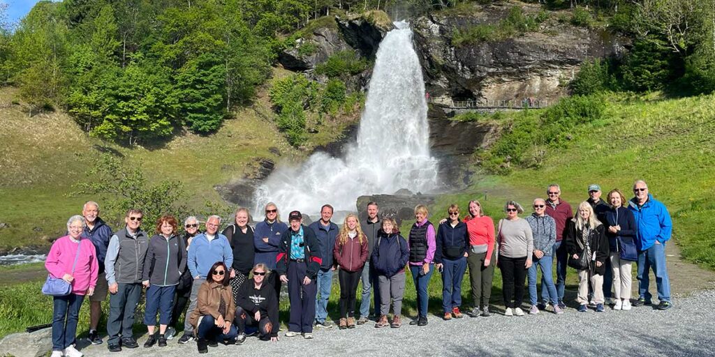 A group of tourists led by a travel director posing for a photo in front of a large waterfall in a sunny, green landscape.