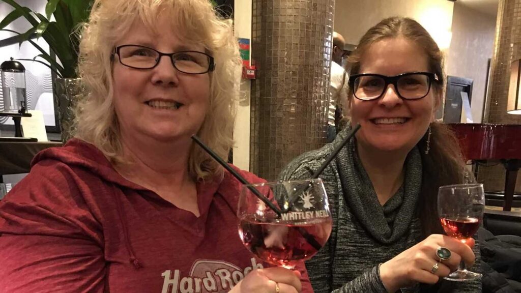 Two women smiling at a table on a Trafalgar tour, each holding a glass of wine, in a cozy indoor setting. One wears a red shirt and glasses, the other a gray scarf