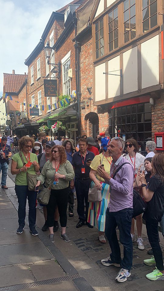 Tourists with a tour guide on a busy, narrow cobblestone street lined with historic half-timbered buildings during their 24 hours in Britain.