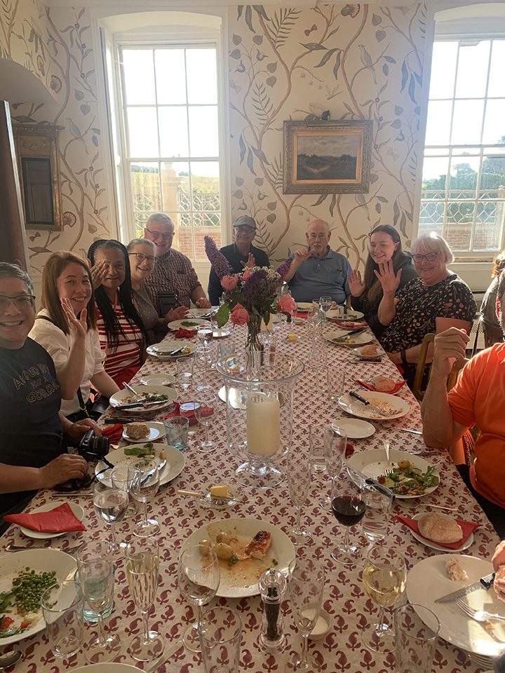 A group of eleven people smiling at a dining table with meals and drinks, in a room with floral wallpaper and natural light, during their 24 hours in Britain.