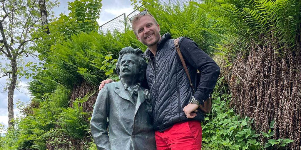 A travel director smiling while standing next to a bronze bust of a historical figure, surrounded by lush greenery.