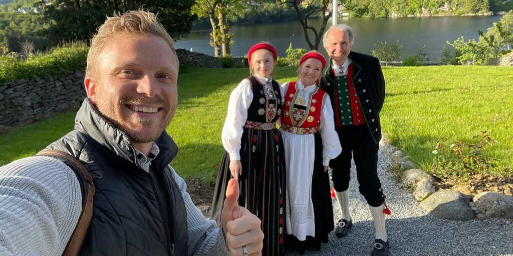 A travel director takes a selfie with three people in traditional Norwegian costumes by a lake, all smiling on a sunny day.