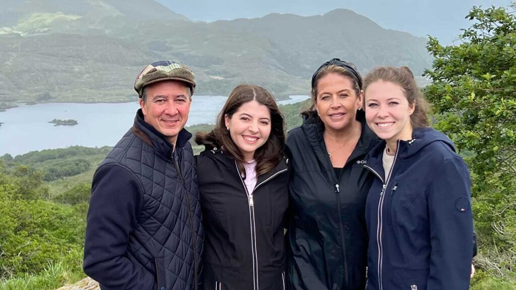 Four people smiling in front of a scenic lakeside view with lush green mountains in the background, celebrating a daughter's graduation.