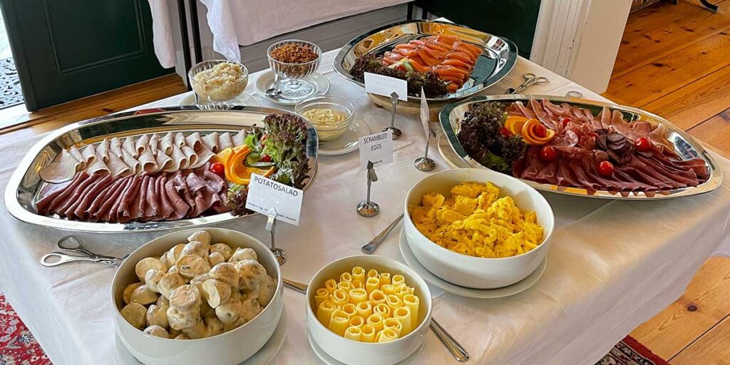 A buffet spread organized by the travel director, with various dishes including cured meats, salmon, and pasta, neatly labeled for guests.