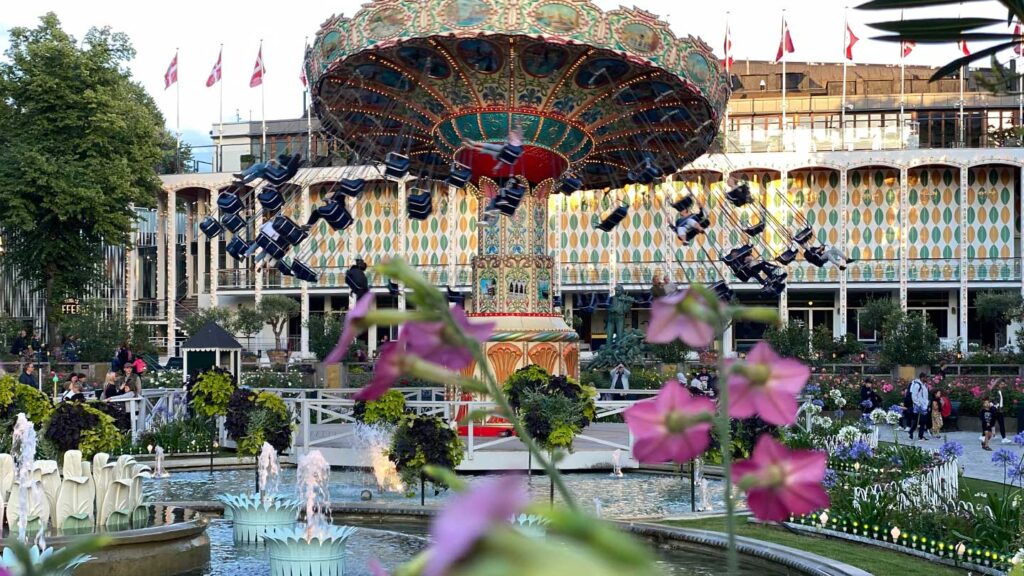 A vibrant carousel with swinging chairs spins in front of a multicolored building, set against a backdrop of international flags and lush garden with fountains, offering visitors a unique way to experience Denmark.