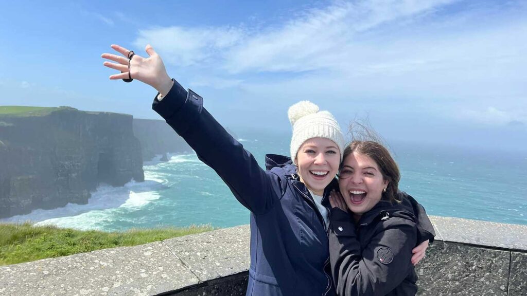 Two joyful women embracing, one raising her arm, at the cliffs of Moher on a windy day with the ocean in the background, celebrating a daughter's graduation.