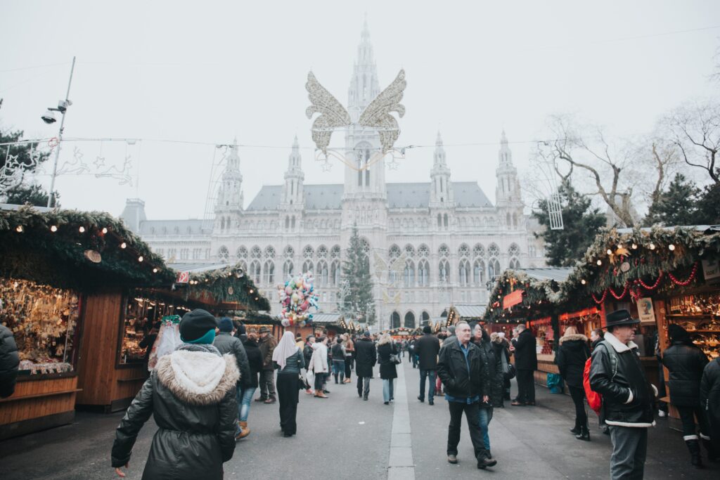Crowded Christmas Markets of Europe in a city square with people walking past festively decorated stalls, large illuminated angel decoration above, and a historic building in the background.