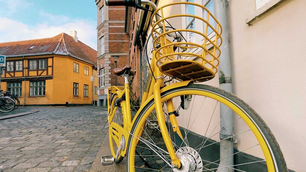 A yellow bicycle with a rear cargo rack parked on a cobblestone street, with traditional Danish buildings in the background.