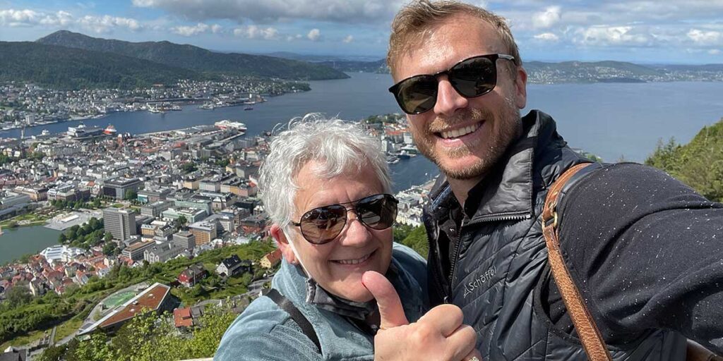 A smiling elderly woman and a young travel director giving a thumbs up, posing for a selfie with a scenic city and fjord in the background.