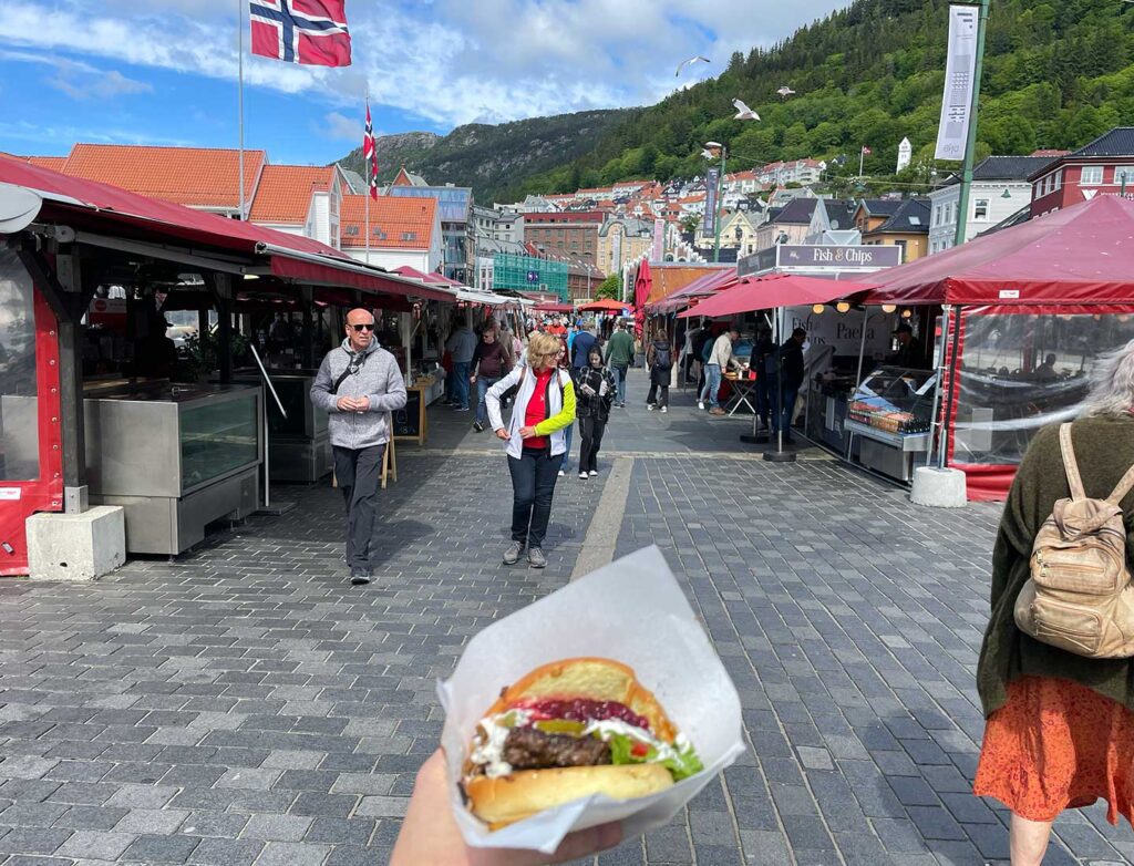 Person holding a hot dog in the foreground with a bustling outdoor market and flag-adorned streets of Bergen, Norway in the background, as viewed by a travel director.