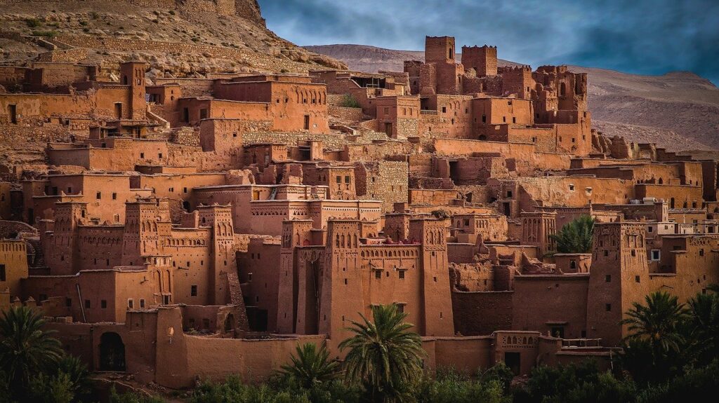 Ancient clay brick buildings of a Moroccan kasbah, surrounded by sparse greenery and set against a hilly backdrop under a cloudy sky, exemplify historical facts about Morocco.