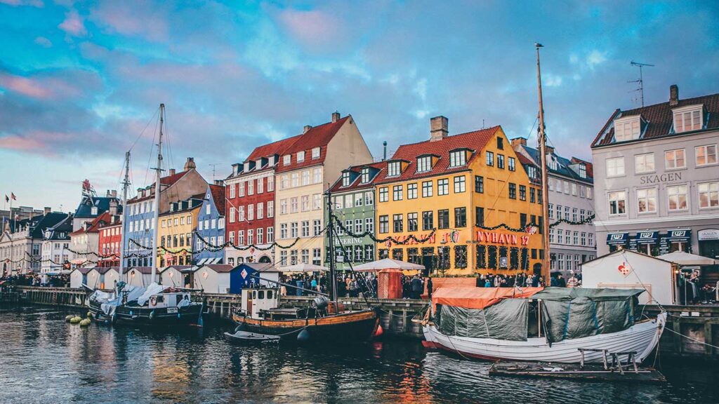 Colorful buildings line the canal at Nyhavn in Copenhagen, Denmark, with boats moored under a vibrant sky at dusk, offering a quintessential experience of Denmark.