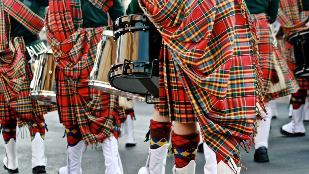 Marching band members in traditional Scottish attire with kilts and knee socks, playing snare drums in a Trafalgar tour parade.