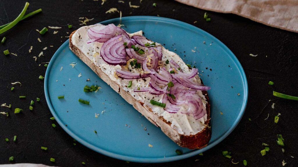 A slice of bread topped with cream cheese, red onion slices, and chopped chives, inviting you to experience Denmark, served on a blue plate.