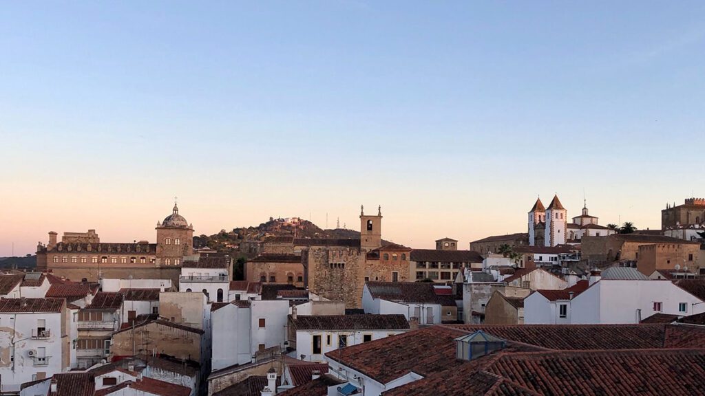 A panoramic view of a historic European town at sunset, showcasing terracotta rooftops and several prominent stone buildings under a soft pink sky, reminiscent of a scene from the House of the Dragon.
