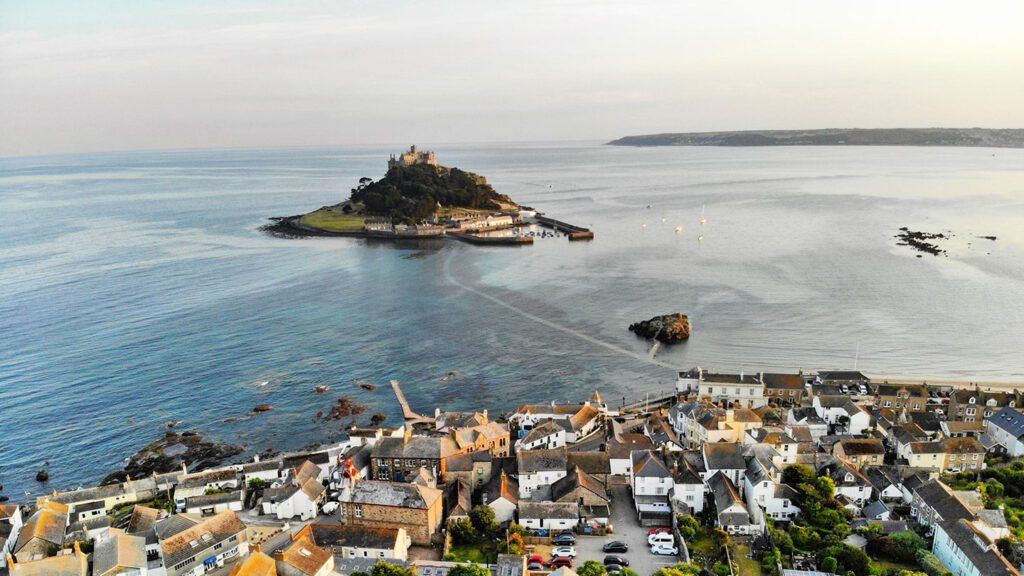 Aerial view of a coastal village with a castle from "House of the Dragon" on a small island, surrounded by the sea and distant hills.