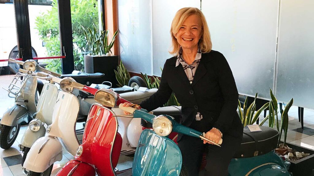 A smiling woman sitting on a blue heritage scooter inside a room with more scooters and plants in the background.