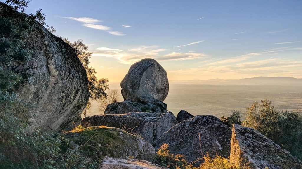 Sunset illuminates a landscape featuring a large, precariously balanced boulder atop a rocky hill with distant mountains and a clear sky, reminiscent of the rugged terrain in "House of the Dragon.