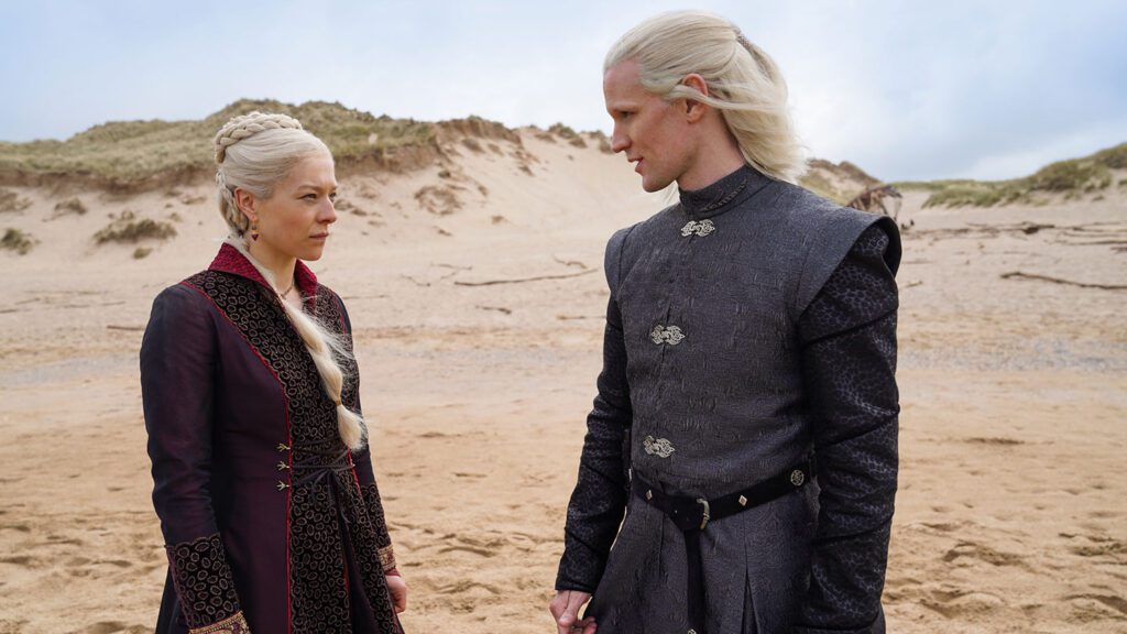 Two characters from "House of the Dragon", a woman and a man, stand on a sandy beach, both dressed in elaborate medieval costumes.