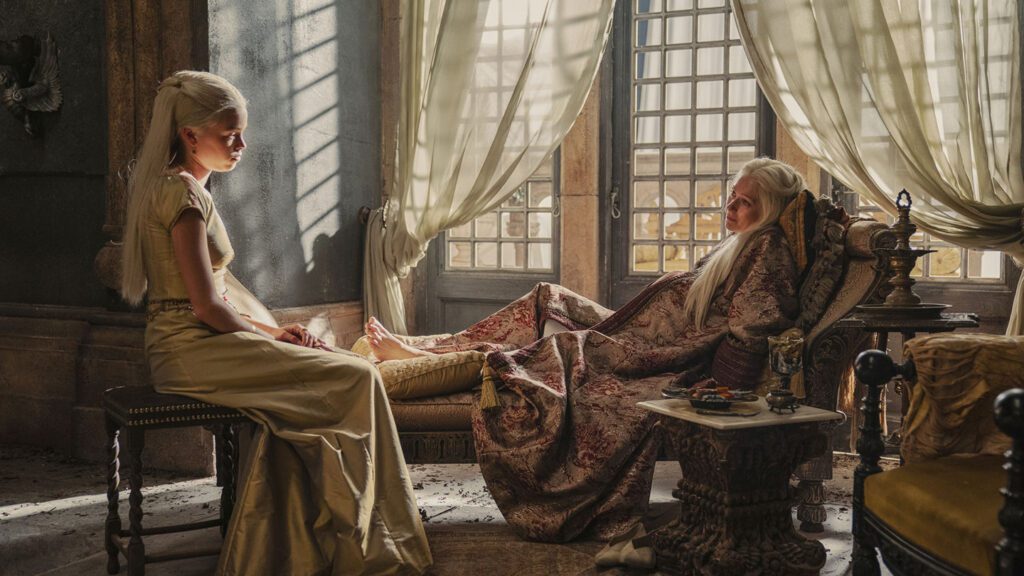 Two women in medieval-style "House of the Dragon" costumes sitting opposite each other in a dimly lit room with flowing curtains and ornate furniture, engaged in conversation.