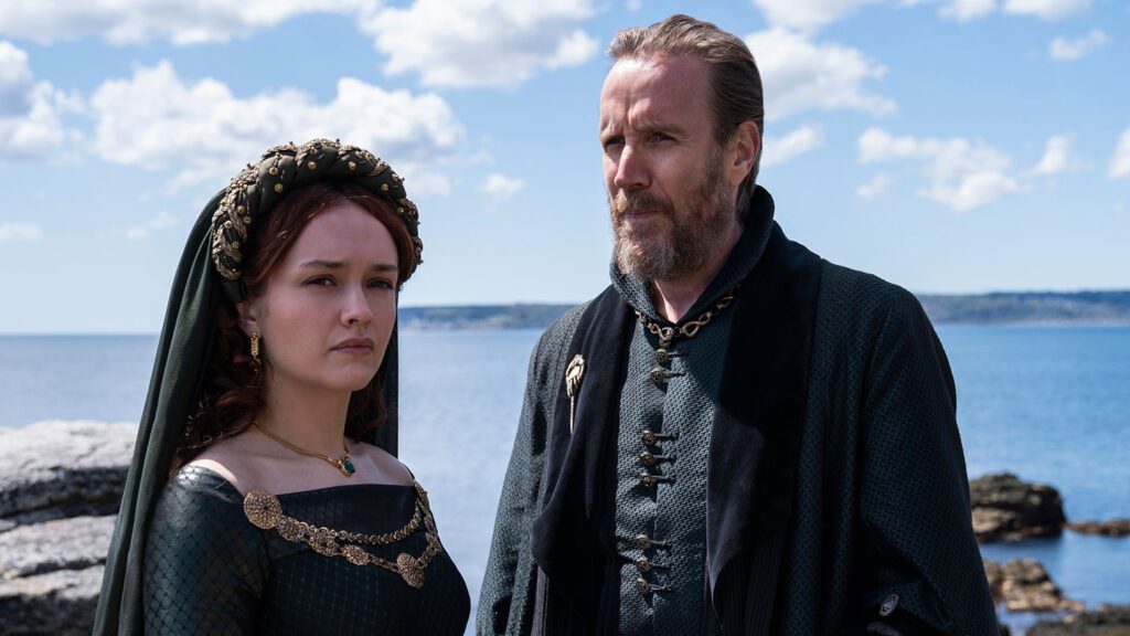 A woman and a man in "House of the Dragon" medieval costumes stand by a coastline under a clear sky.