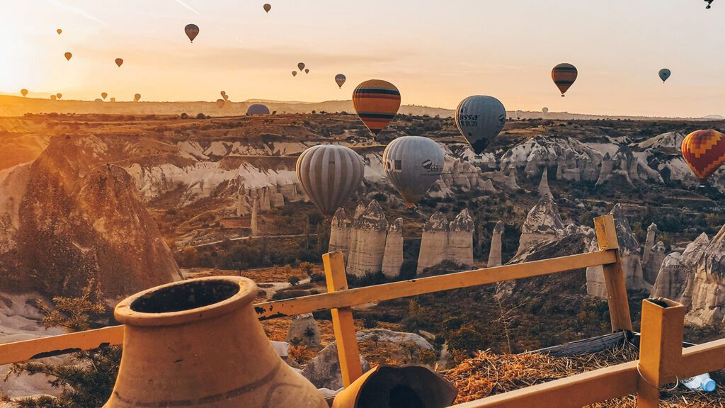 Hot air balloons inspire kids to travel as they float above a rocky landscape at sunrise, framed by a wooden railing and clay pots in the foreground.