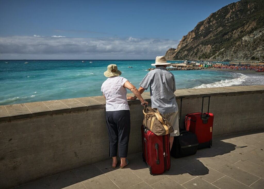 Two senior adults standing by a seaside railing, overlooking a beach, each with a red suitcase, discussing how not to lose luggage on a partially sunny day.