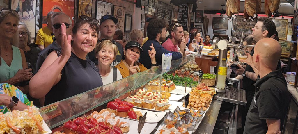 A bustling tapas bar with patrons and a server, displaying an array of colorful tapas in the foreground.