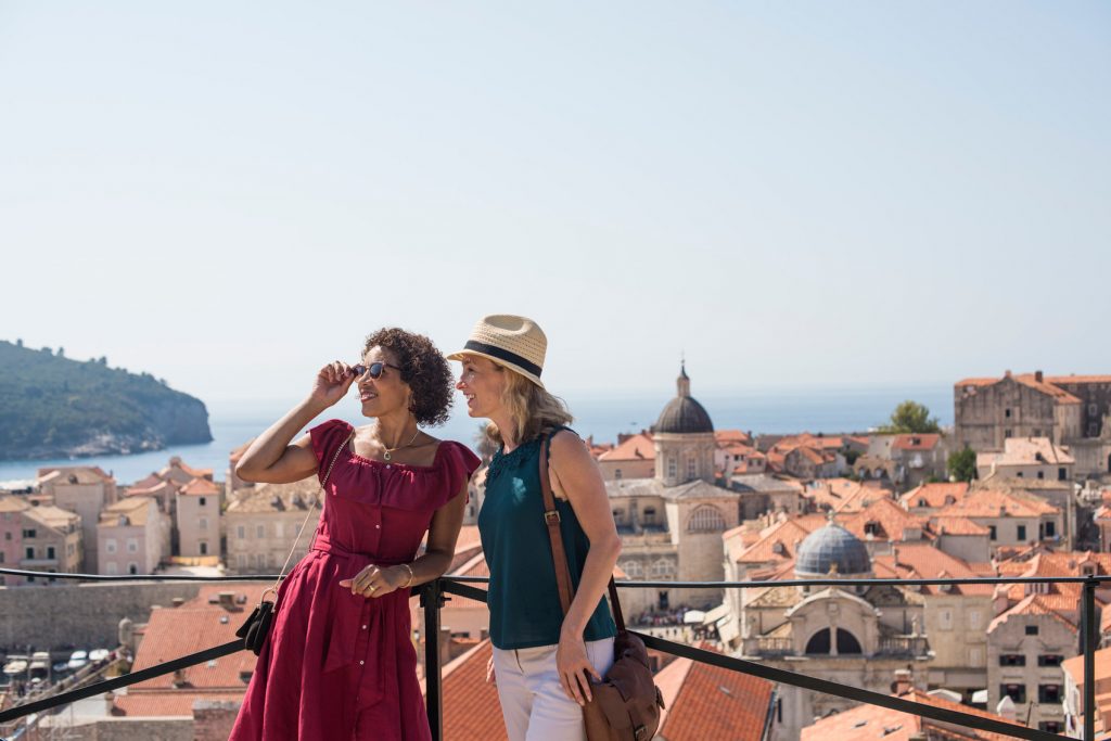 Two women enjoying a scenic view of their favourite travel destination, a coastal city from a high vantage point, one pointing at the distance, both wearing summer outfits and hats.