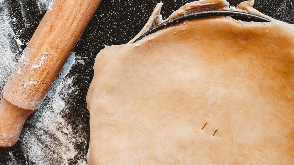 Rolled out dough for a pie recipe with a wooden rolling pin and a wire dough cutter on a floured surface.
