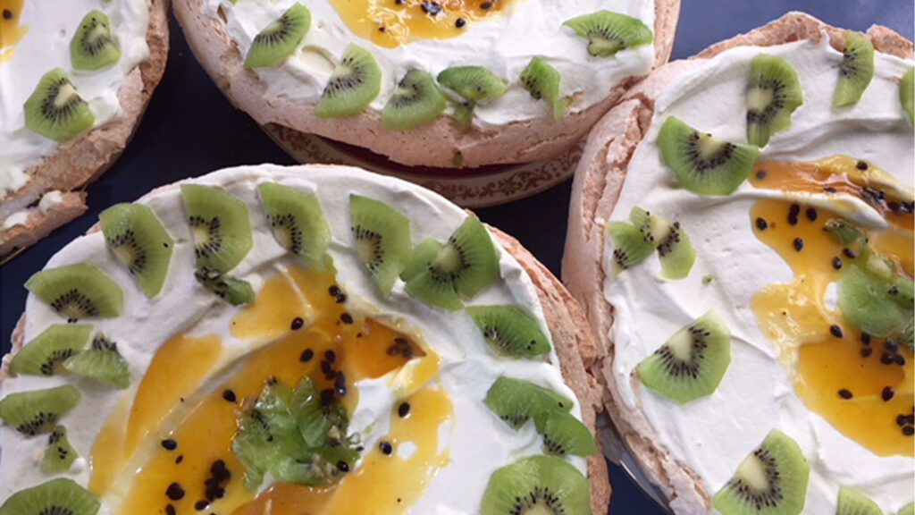 Four New Zealand pavlova desserts topped with whipped cream, kiwi slices, and passion fruit pulp on a dark surface.
