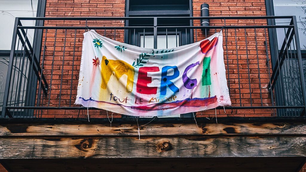 Colorful homemade "how to say 'thank you'" banner with artistic decorations hanging from a balcony railing on a brick building.