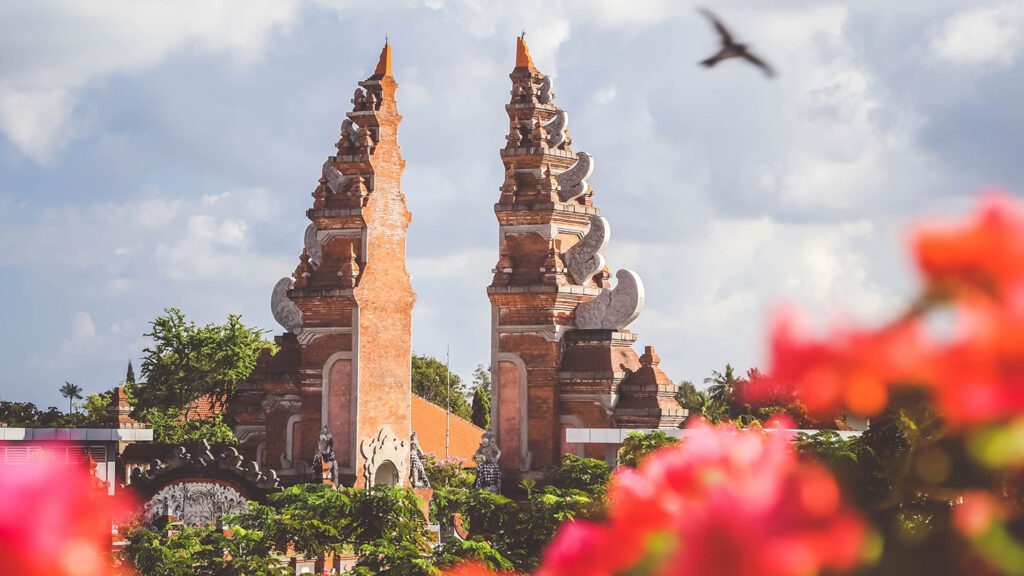 Twin ornate terracotta spires of a traditional temple rise against a cloudy sky, framed by out-of-focus pink flowers in the foreground and how to say 'thank you' in