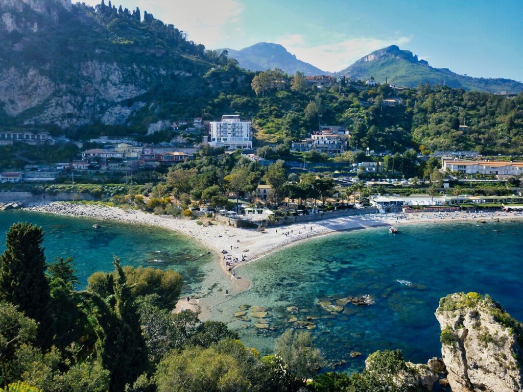 Aerial view of a curved beach with clear turquoise waters, surrounded by green hills and a small town in Sicily, under a clear blue sky.